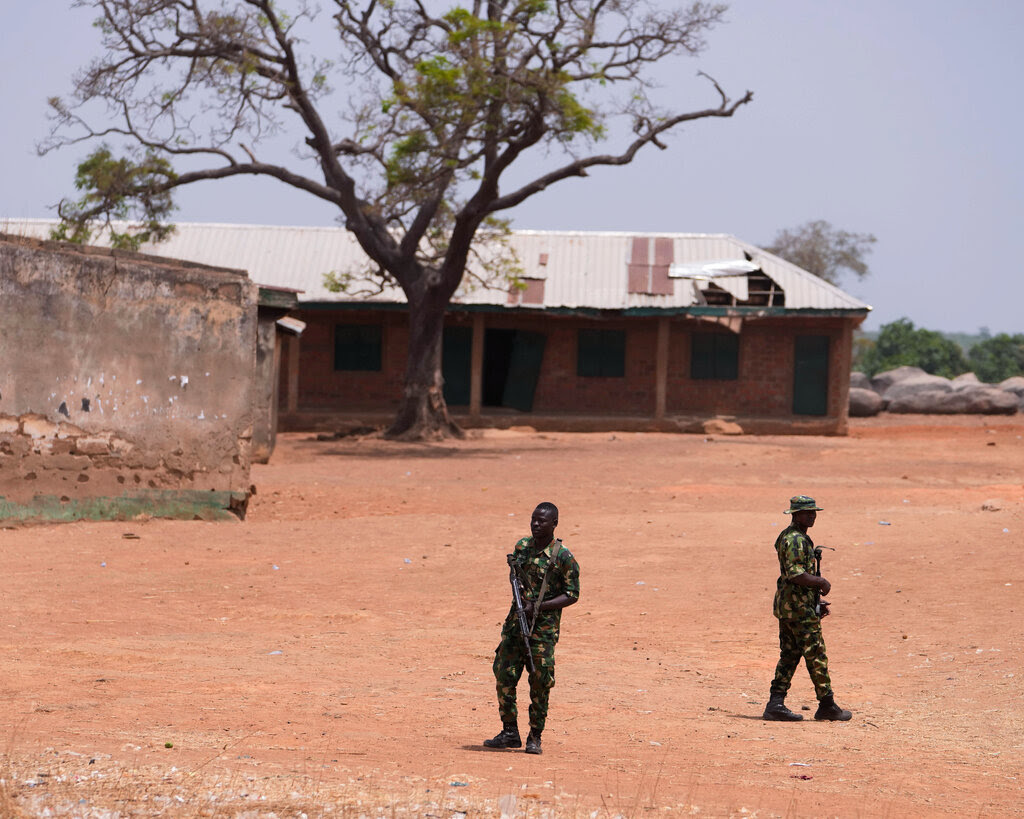 Two people in military attire stand in a dirt field outside a dilapidated building.