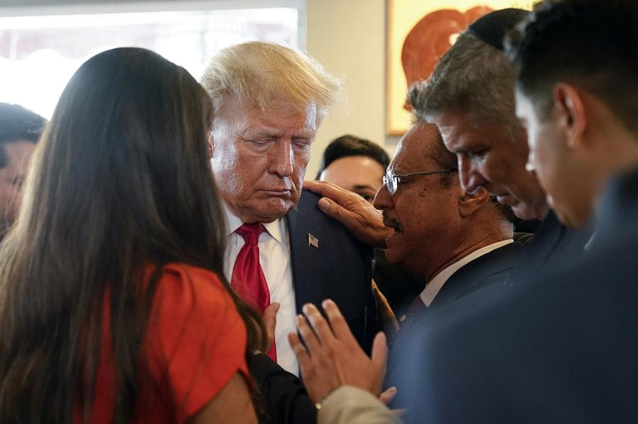 Former President Donald Trump prays with Mario Bramnick, a pastor. There are other people gathered in prayer together.