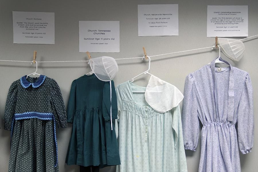 Dresses donated by sexual assault survivors from Amish and other plain-dressing religious groups hang on a clothesline beneath a description of each survivors' age and church affiliation.