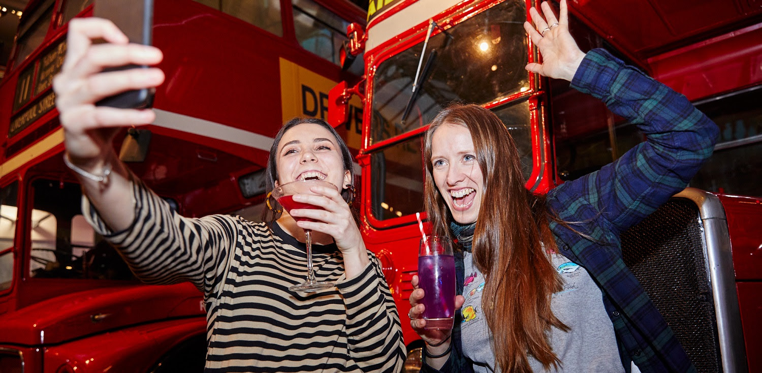 Two young people holding cocktails and taking a selfie in front of historic red London buses