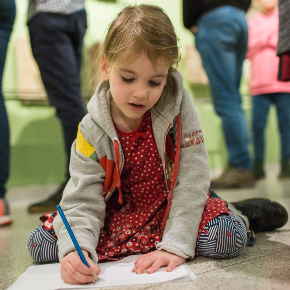 A young girl kneels on the floor of the Museum and draws on a piece of paper