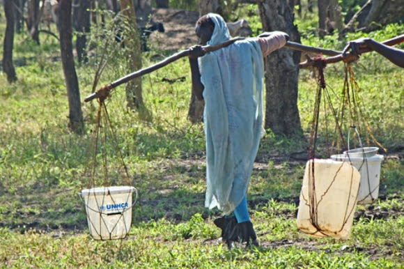 Una refugiada de Sudán recorre un largo camino desde el punto de abastecimiento de agua más cercano a su alojamiento temporal, cargada con recipientes de plástico llenos de agua. El CICR está mejorando la red de abastecimiento de agua para hacer más liviano su trabajo y el de tantas otras.