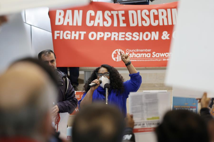A woman speaks at a mircrophone at a rally in front of a sign reading "BAN CASTE DISCRIMINATON"