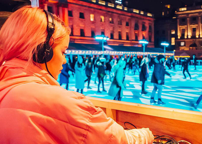 A women is DJing on rink-side decks beside the ice rink at Somerset House. The ice rink is lit a vivid blue with people zooming around in the background.