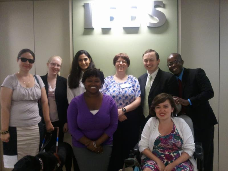 A group of eight people in business suits blouses or dresses smile at the camera One has a guide dog another is seated in the wheelchair On the wall behind them is the logo for the International Federation Electoral Systems IFES