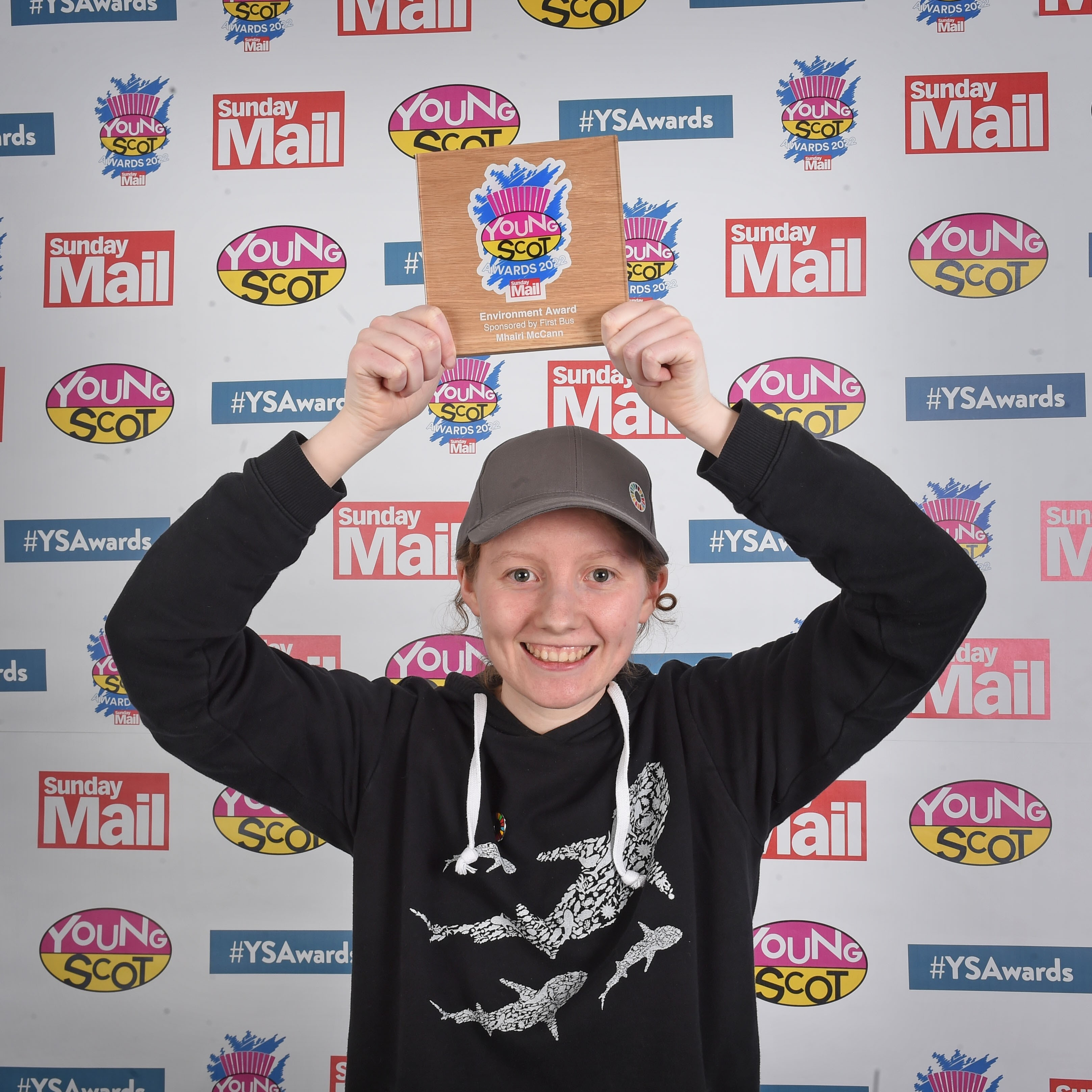 Mhairi wearing black hoodie with whales on it and grey cap.  Mhairi is smiling and holding Young Scot Award above head.  Background is a logo wall of Young Scot, Sunday Mail and Young Scot Awards.