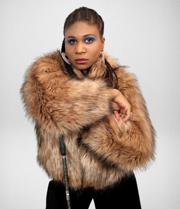 Lachi is a blind, black woman wearing a brown fuzzy fur jacket, large circle earings and stylish blue eyeshadow withlong braids