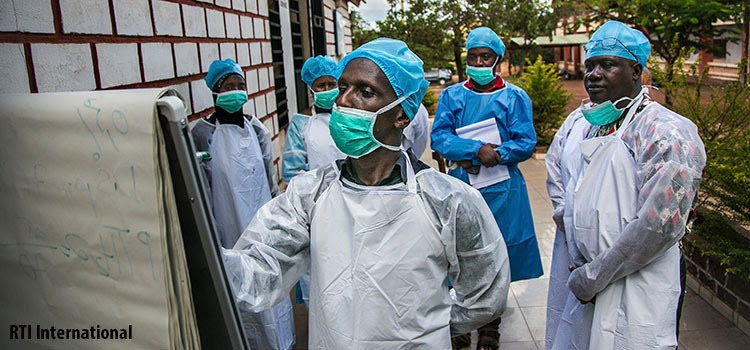 Local healthcare workers in Guinea wearing face masks and medical gear with a man in front of the group writing on a paper board as other colleagues in the back pay attention. Photo: RTI International