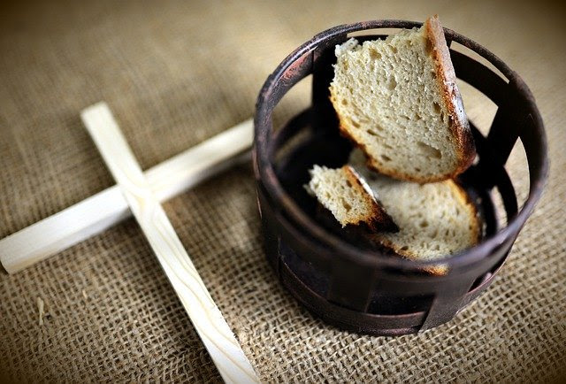 A crucifix made of pale wood lies next to a rustic metal basket filled with chunks of bread.