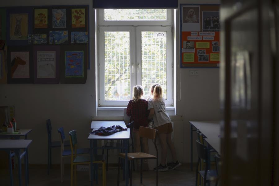 Two young girls stand together in front of the window in their classroom.