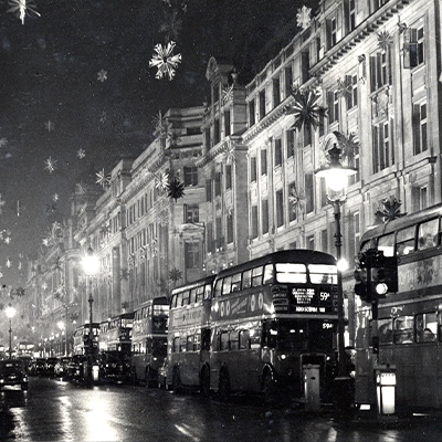 Historical photo showing buses travelling through a city street at night with snowflake decorations displayed 