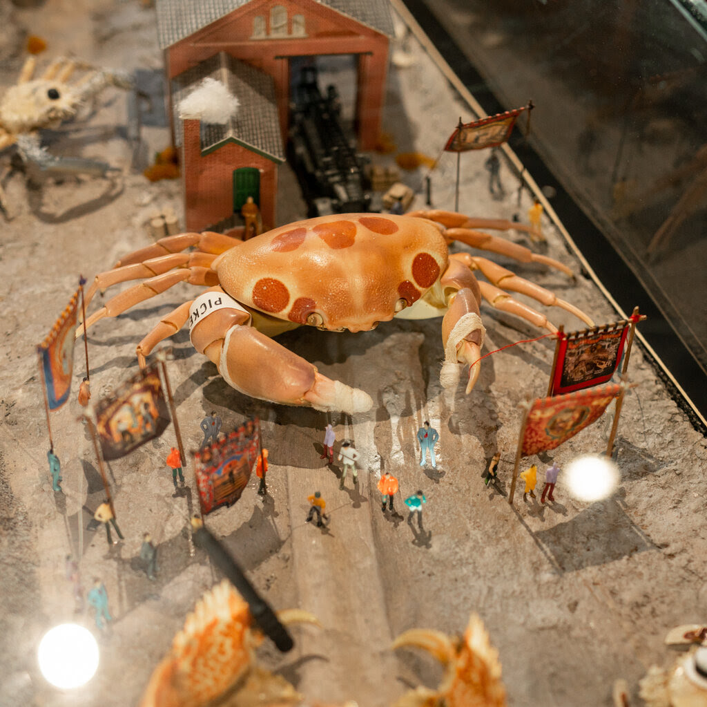 In a glass case, a model crab towers over models of people and a train. 