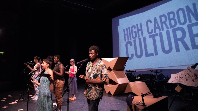 A group of people performing in a dark space with microphones in front of them. On the wall is a projected image of a white card with the words "HIGH CARBON CULTURE".