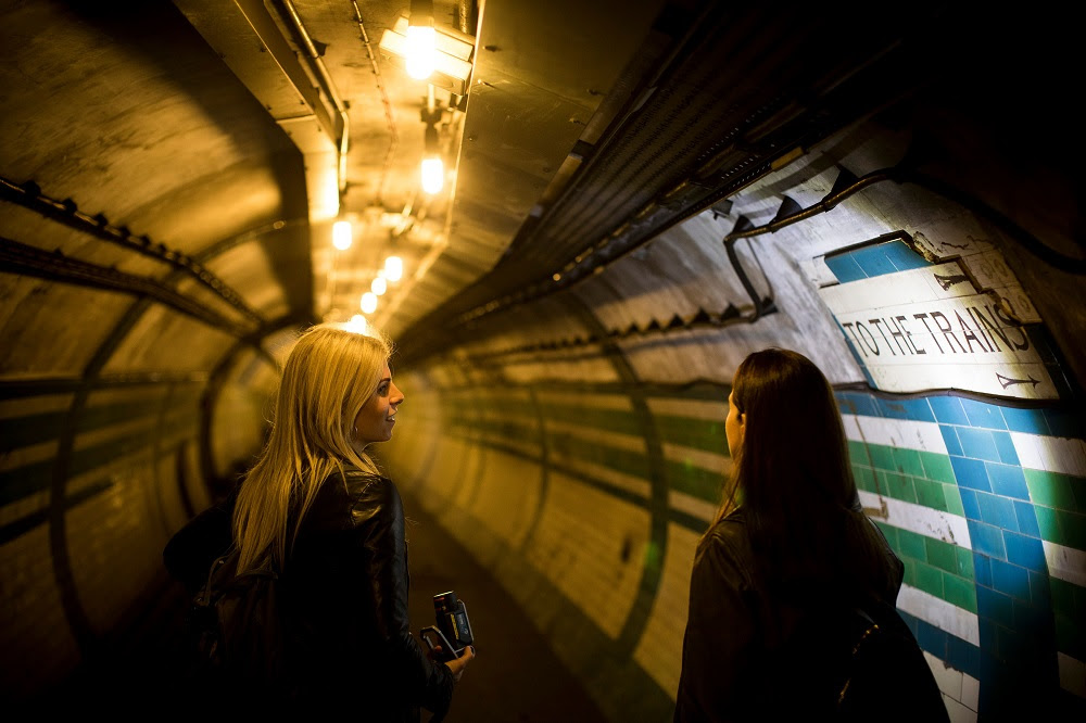 Two people in a tiled tunnel shining torch on sign reading To the trains