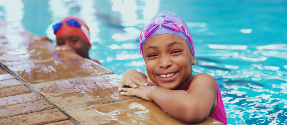 Two girls in a pool, holding on to the side of the pool. One girl with goggles and swim cap, smiling.