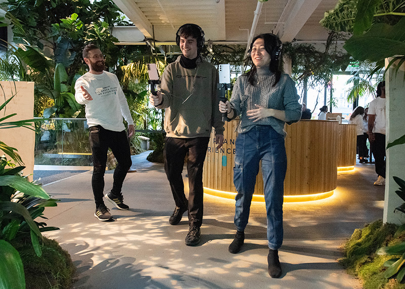 A photo of two industry placement participants working. They are on set testing light levels in a room surrounded by plants.