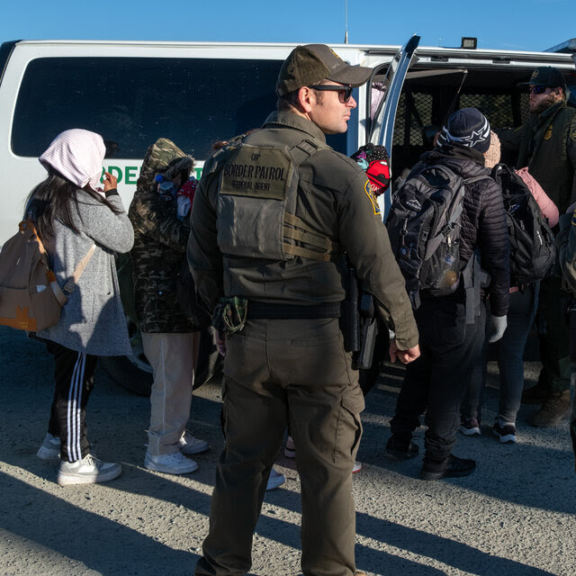Border Patrol officers watch as people carrying backpacks enter a white van.