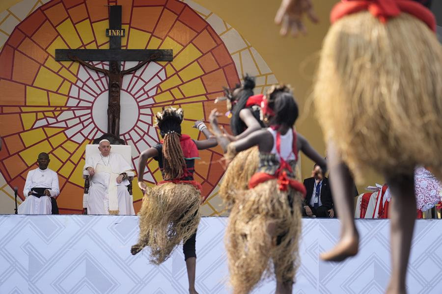 Pope Francis watches traditional dancers perform at Martyrs' Stadium In Kinshasa, Democratic Republic of Congo. He is sitting in front of a large crucifix with a brightly colored mural behind it.