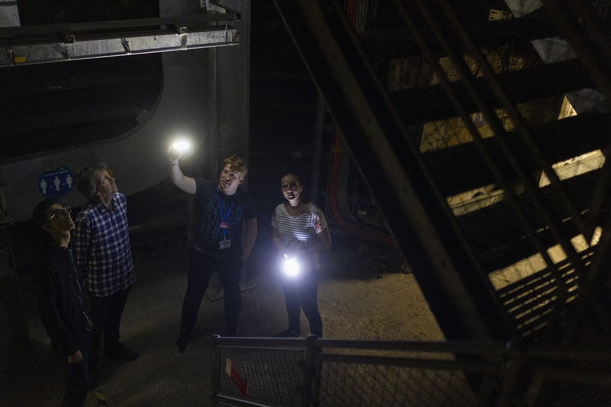 Four people holding torches looking up an underground metal staircase