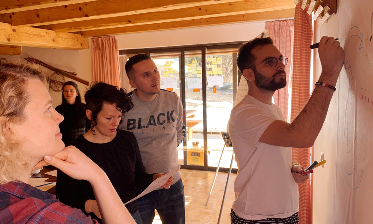 A man with light skin with olive undertones writes on a whiteboard as a small group of people with light skin look on with thoughtful expressions, some resting their hand on their chin, others looking at notes.