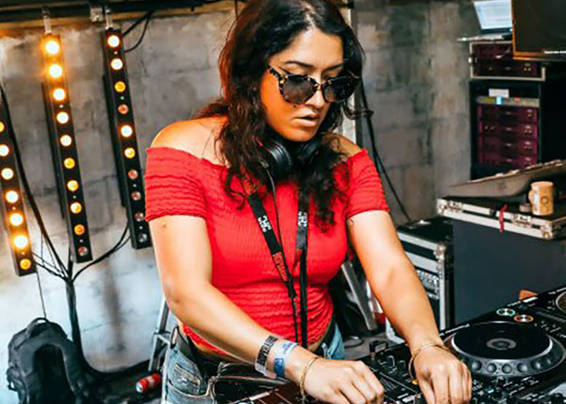 A photo of DJ Manara on the decks. They have shoulder length brown hair, are wearing sunglasses and a vibrant red top.