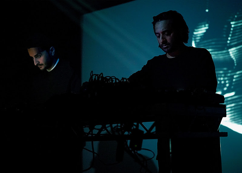 A photo of Imran Perretta and Paul Purgas performing as AMRA. They are stood behind electronic sound equipment and behind them is a projection of grey-blue and light.