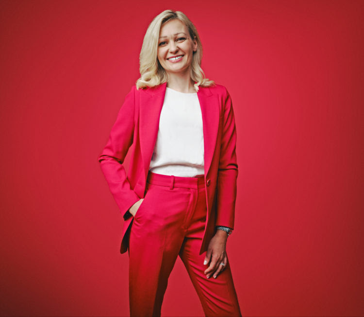 Image shows a woman dressed in a red suit against a red background. She has blonde hair and has one hand in her pocket with a smile on her face.