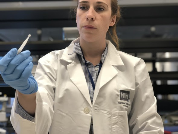 A woman in a white lab coat uses her gloved hand to hold up a small white blood microsampling device.