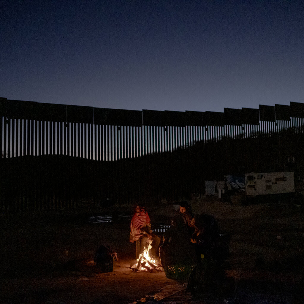 A group of people wearing blankets huddling around a fire. The border wall extends past them in the background.