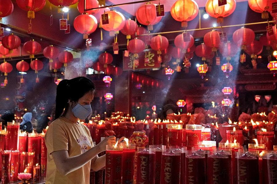 A woman lights a candle during lunar New Year celebrations. There are many rows of red candles that are already lit behind her. There are also red paper lanterns hanging overhead.