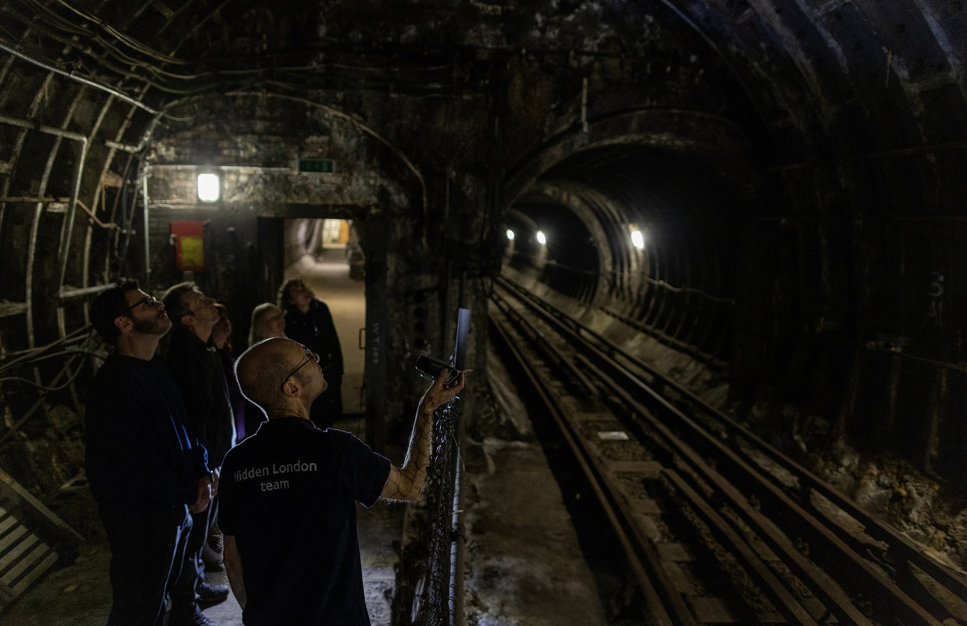 Tour guide shining torch into black-walled Underground tunnel as tour group look along the train tracks