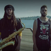 Two men stand on a pier near a lake, one has a sax around his neck.