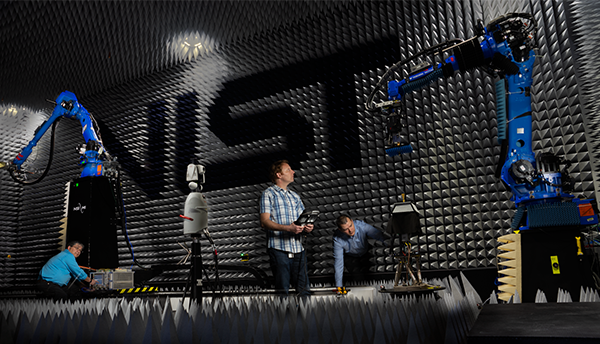 Researchers in large anechoic chamber (antenna calibration facility) with NIST logo on wall looking at robotic arms and equipment.