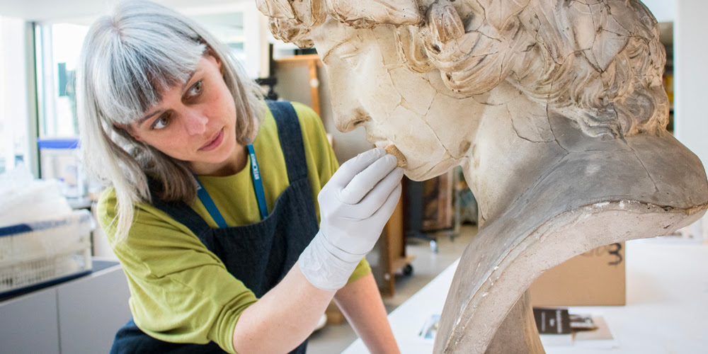 A conservator cleans a large cast bust of the head of a man